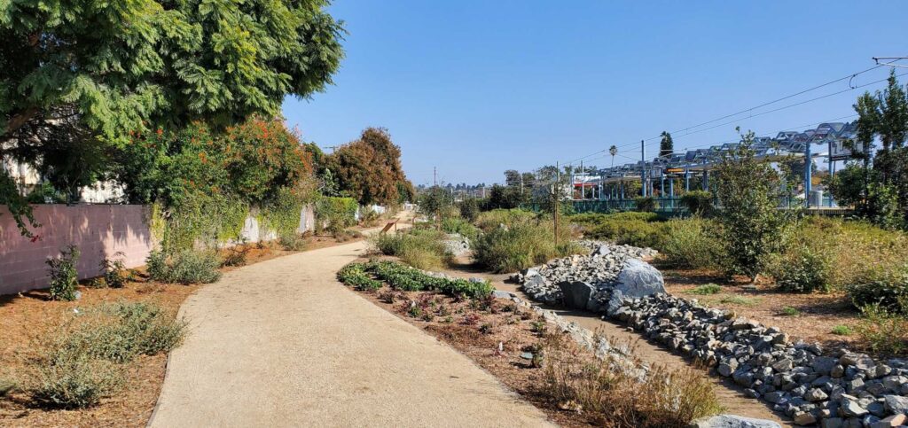 A sunny park pathway with greenery and rocks under a clear blue sky.