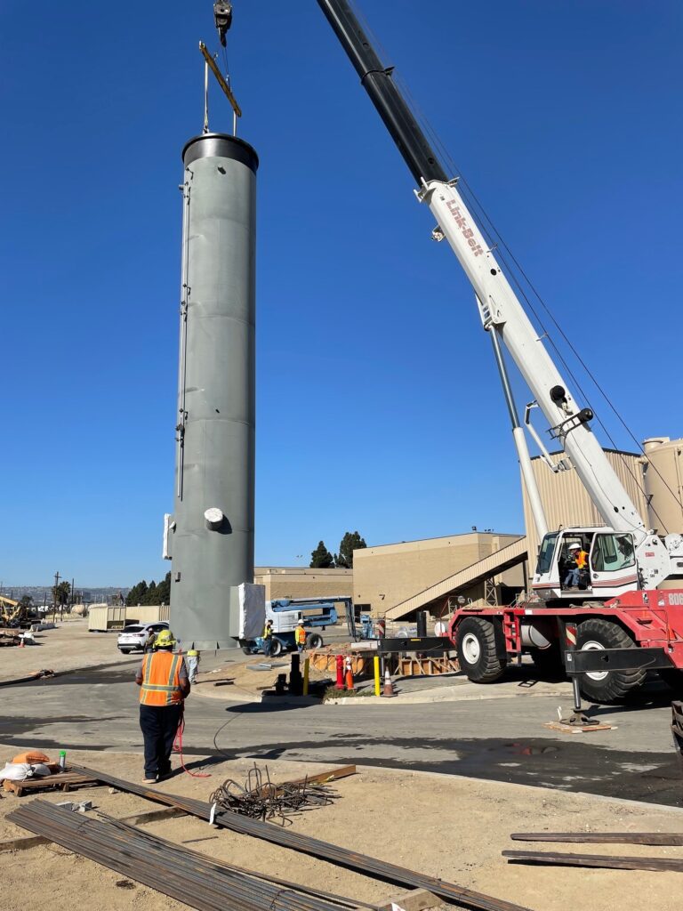 Construction workers operate a crane near a tall cylindrical structure under a clear blue sky.