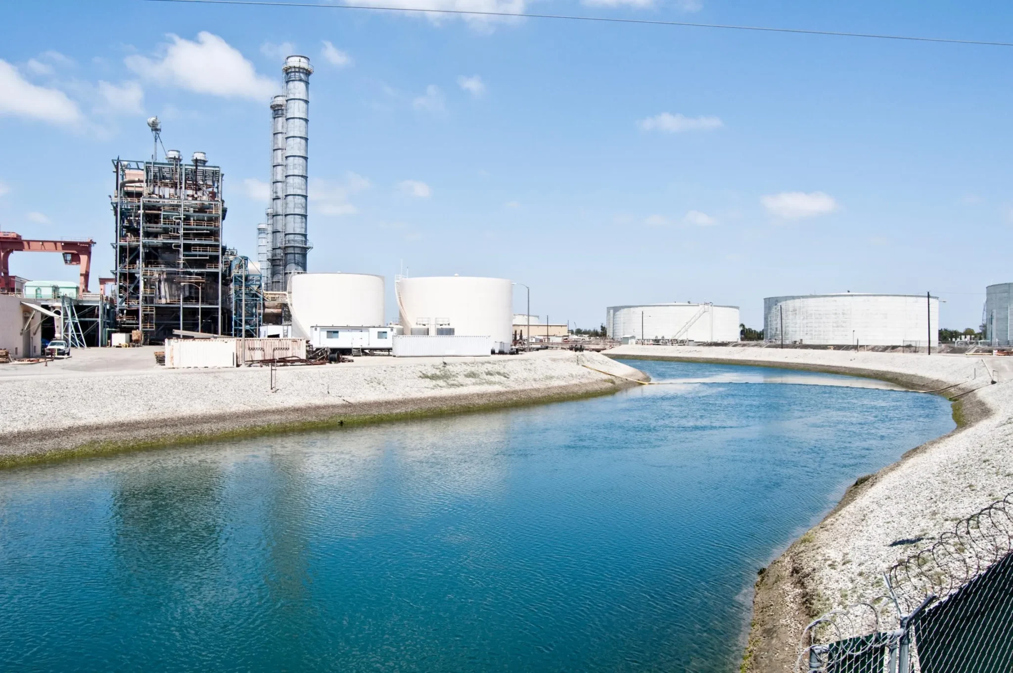Industrial storage tanks beside a water canal under a clear sky.