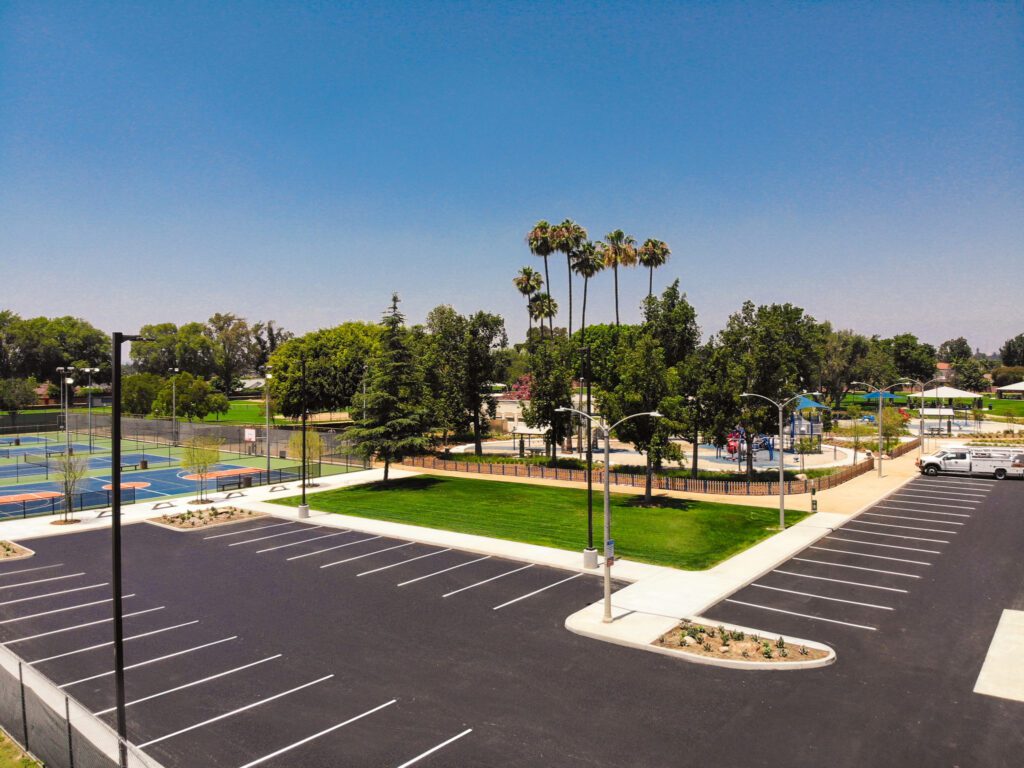 Empty parking lot next to a green park with palm trees under a clear blue sky.