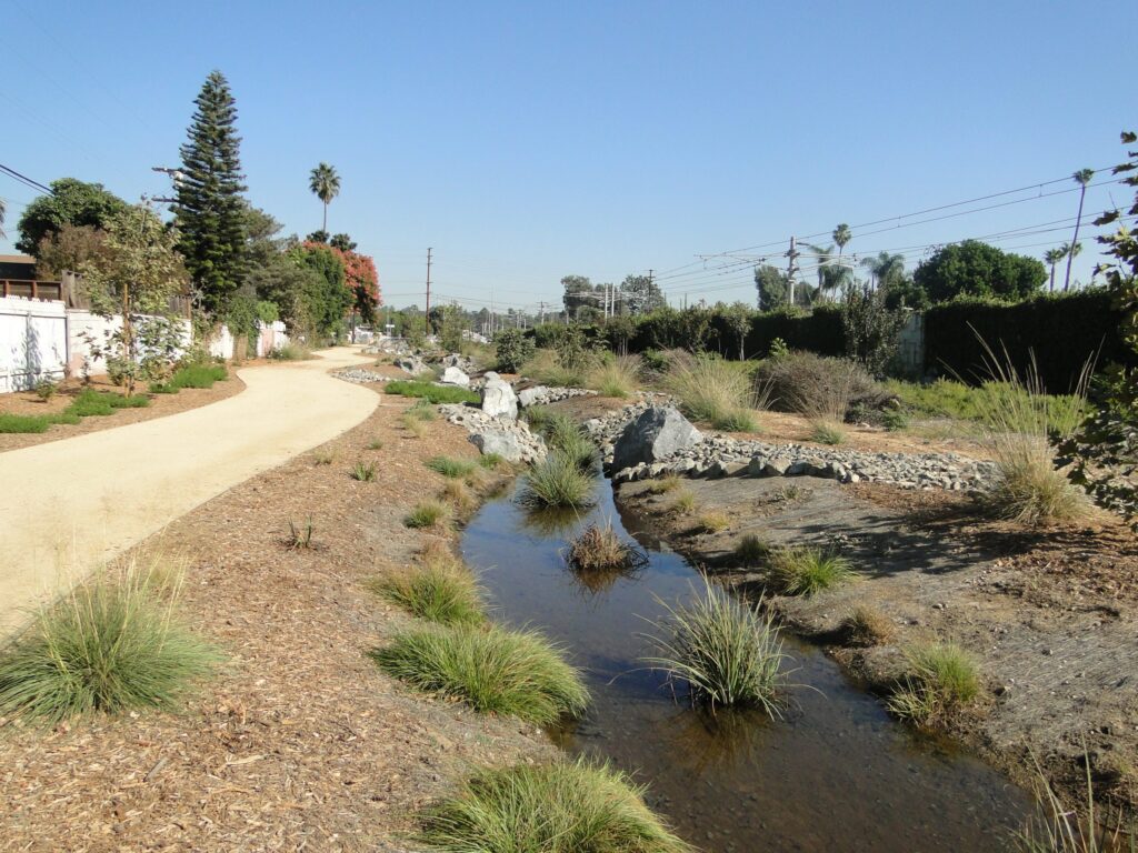 A small stream flows alongside a dirt path with greenery and trees under a clear sky.