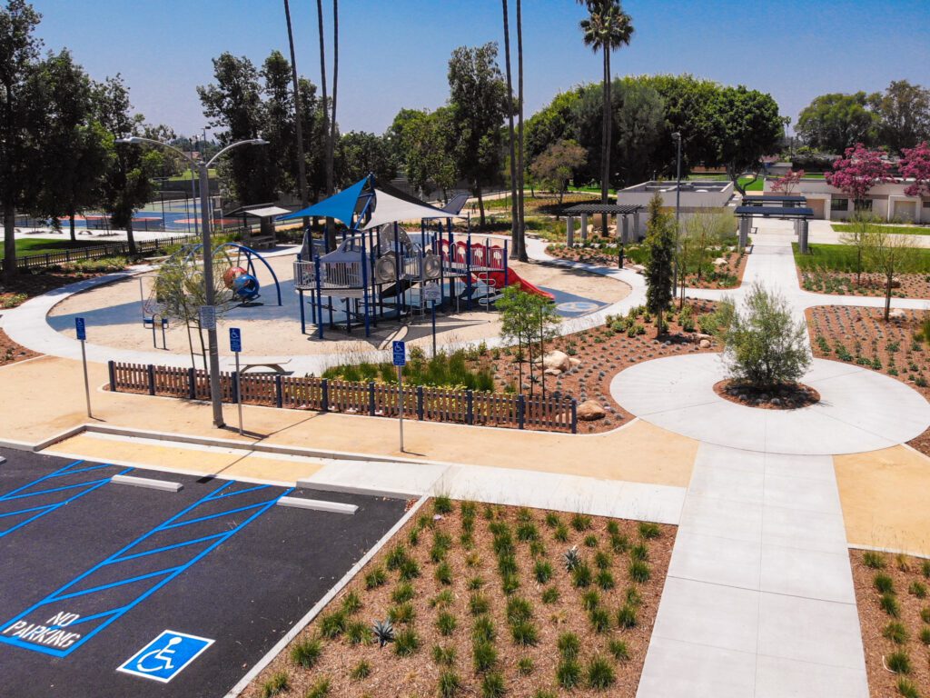 A sunny playground with slides and climbing structures near a parking lot.