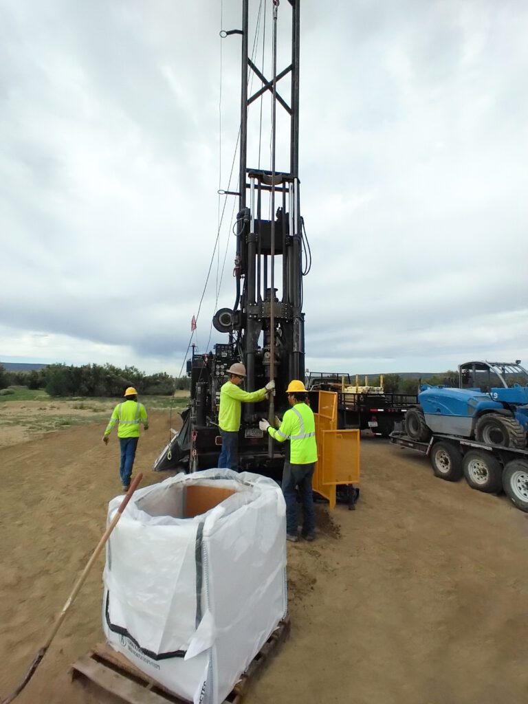 Workers operate a drilling machine at a construction site under a cloudy sky.