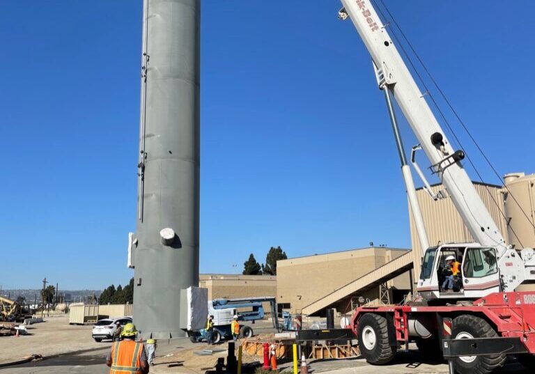 Construction workers operate a crane near a tall cylindrical structure under a clear blue sky.