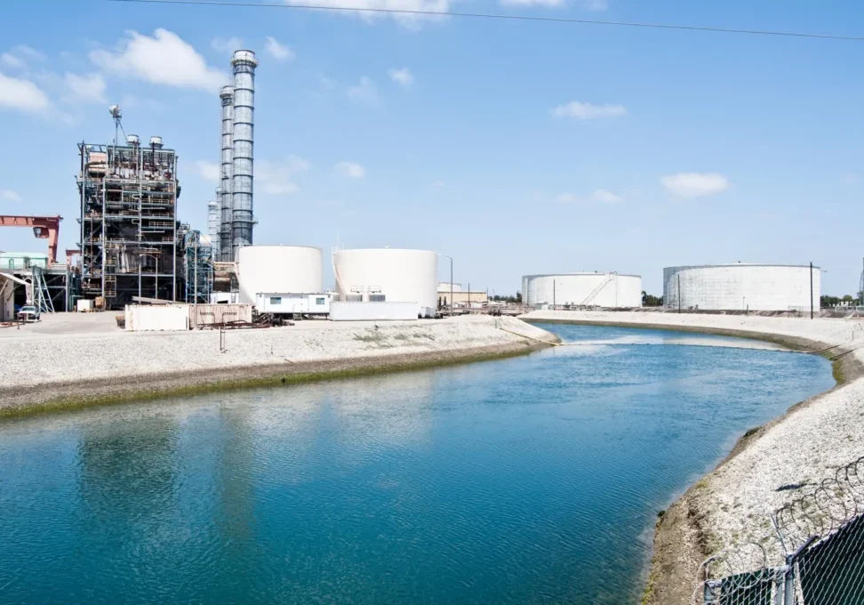 Industrial storage tanks beside a water canal under a clear sky.